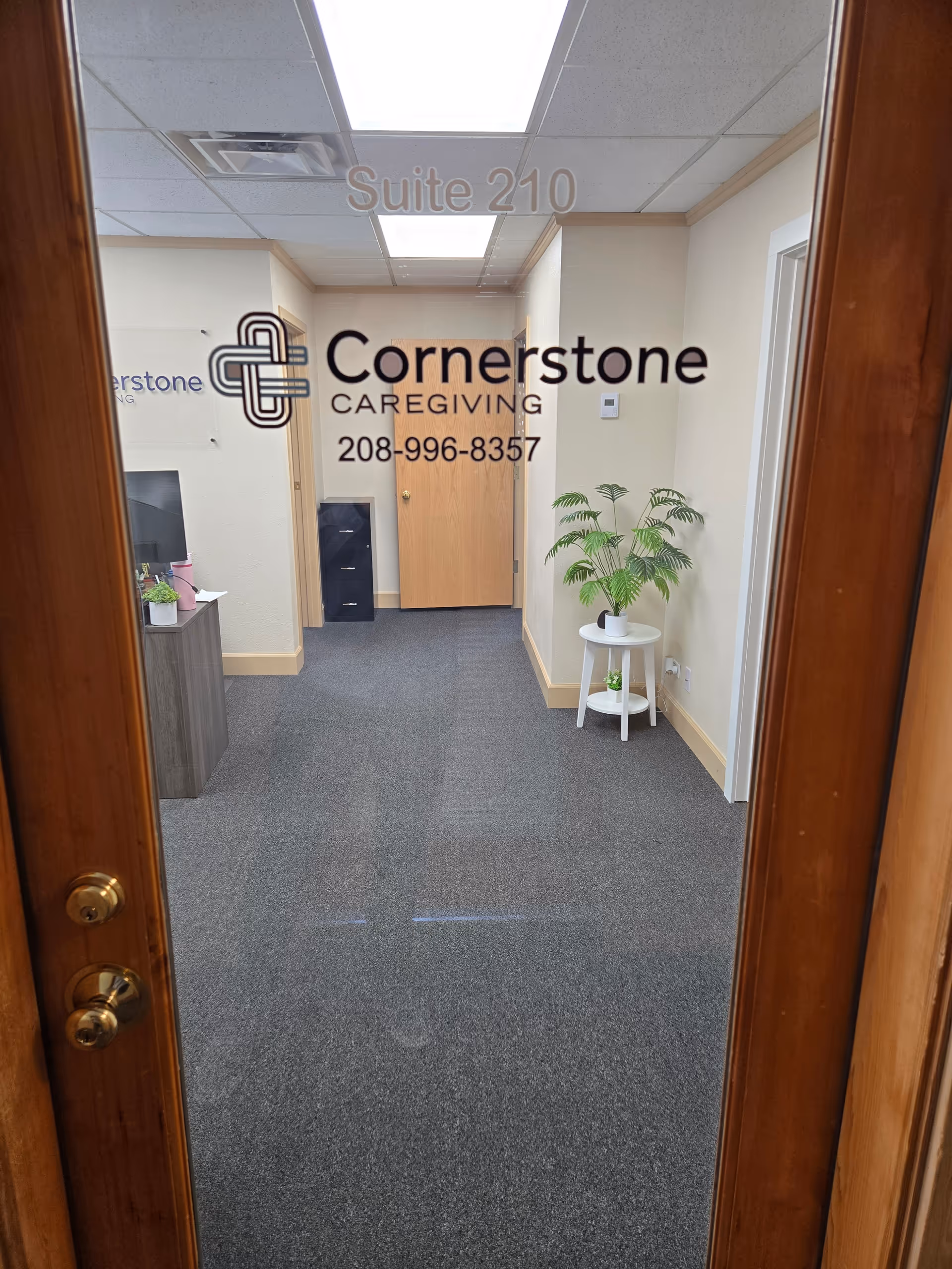 Interior hallway with glass door displaying Suite 210 and Cornerstone Caregiving, plant and furniture visible.