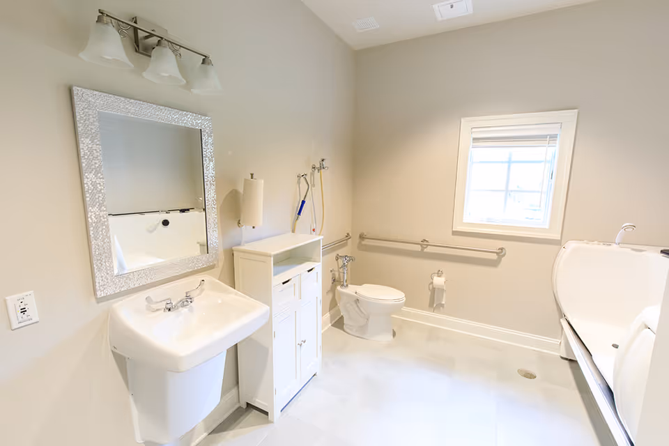 Bright senior bathroom with sink, mirror, toilet, grab bars, and a walk-in tub.