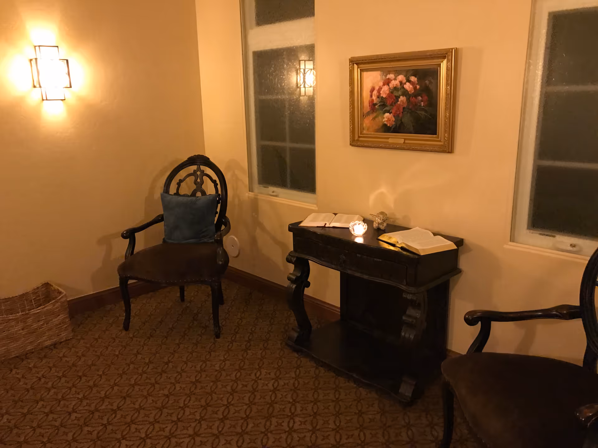 Warm, dim lounge with two chairs, a dark console table, books, a painting, and windows.
