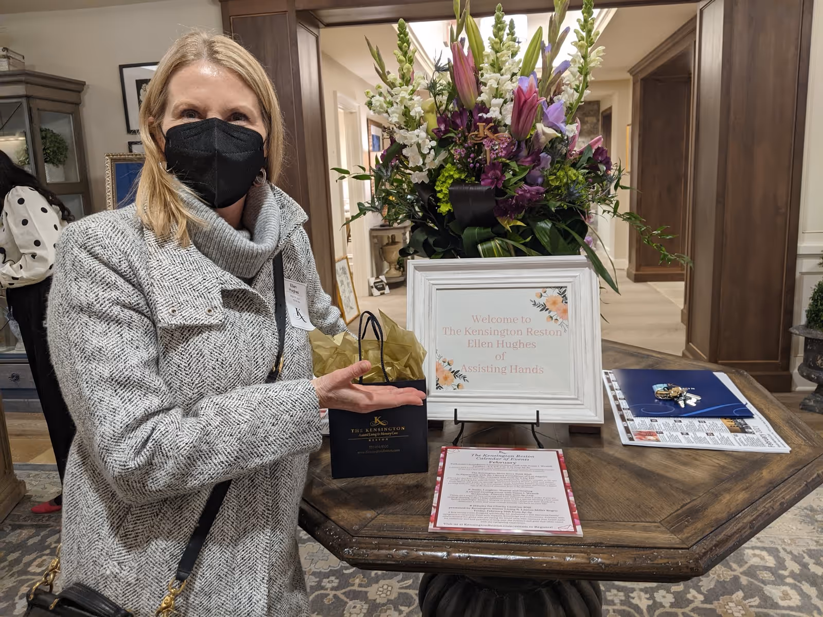 Staff member in a mask hands a small gift beside a floral welcome display.