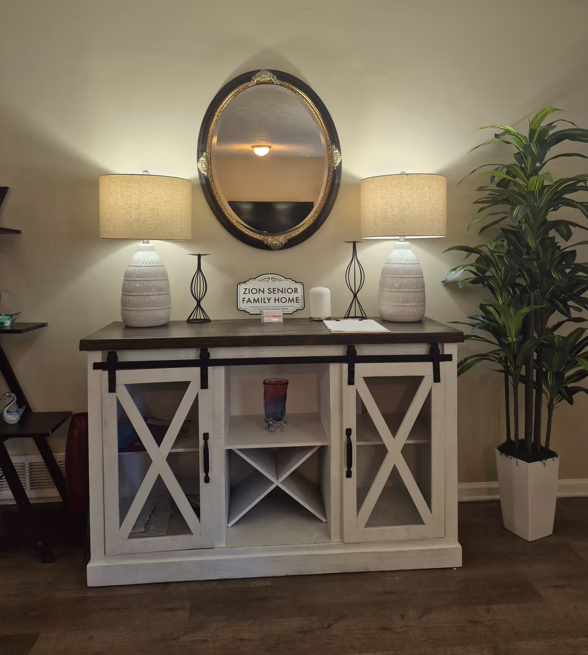 A white console table with lamps, mirror, sign, and a tall plant in a neutral lobby.
