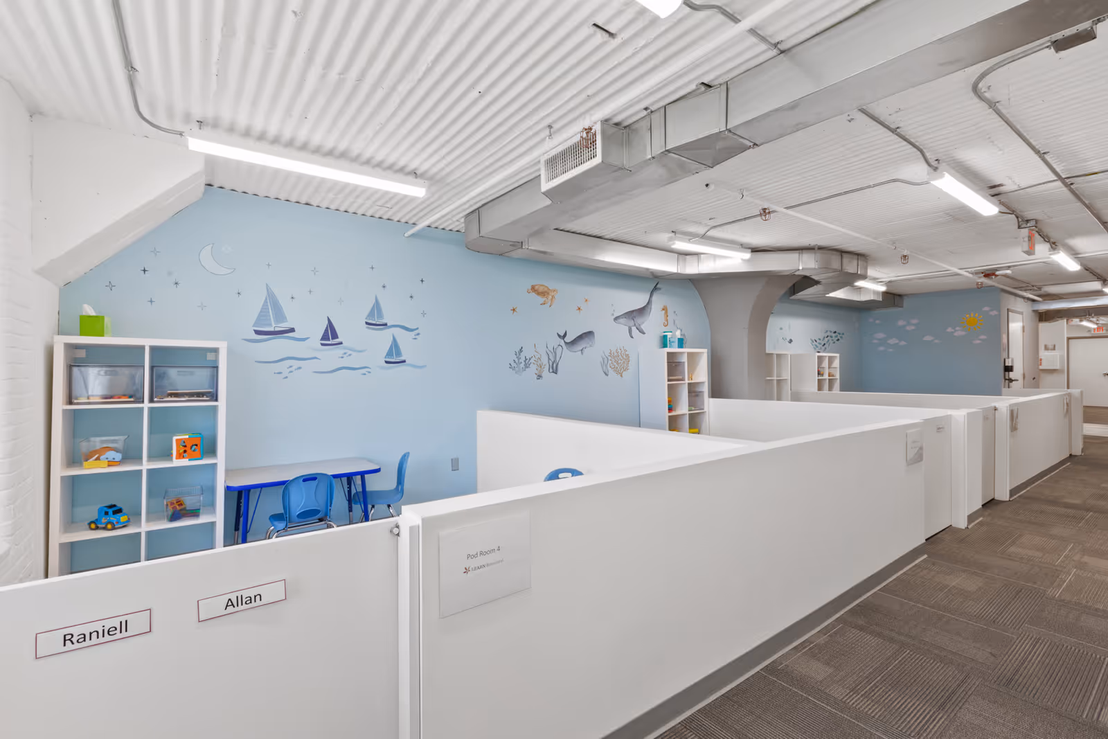 Bright airy hallway with white partition cubicles, blue mural walls, and small activity tables.