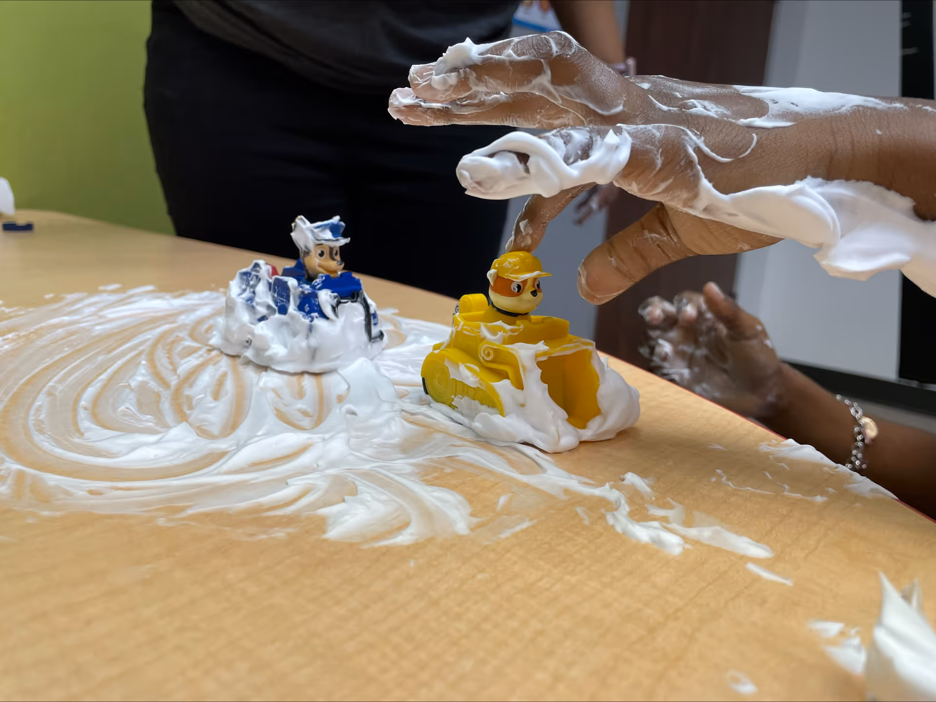 Hands mold white foam around toy construction vehicles on a wooden table.
