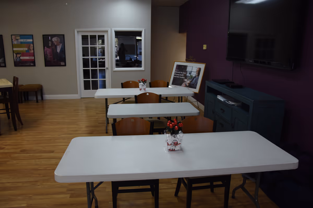 Senior living facility dining area with white folding tables, chairs, and a wall-mounted TV.