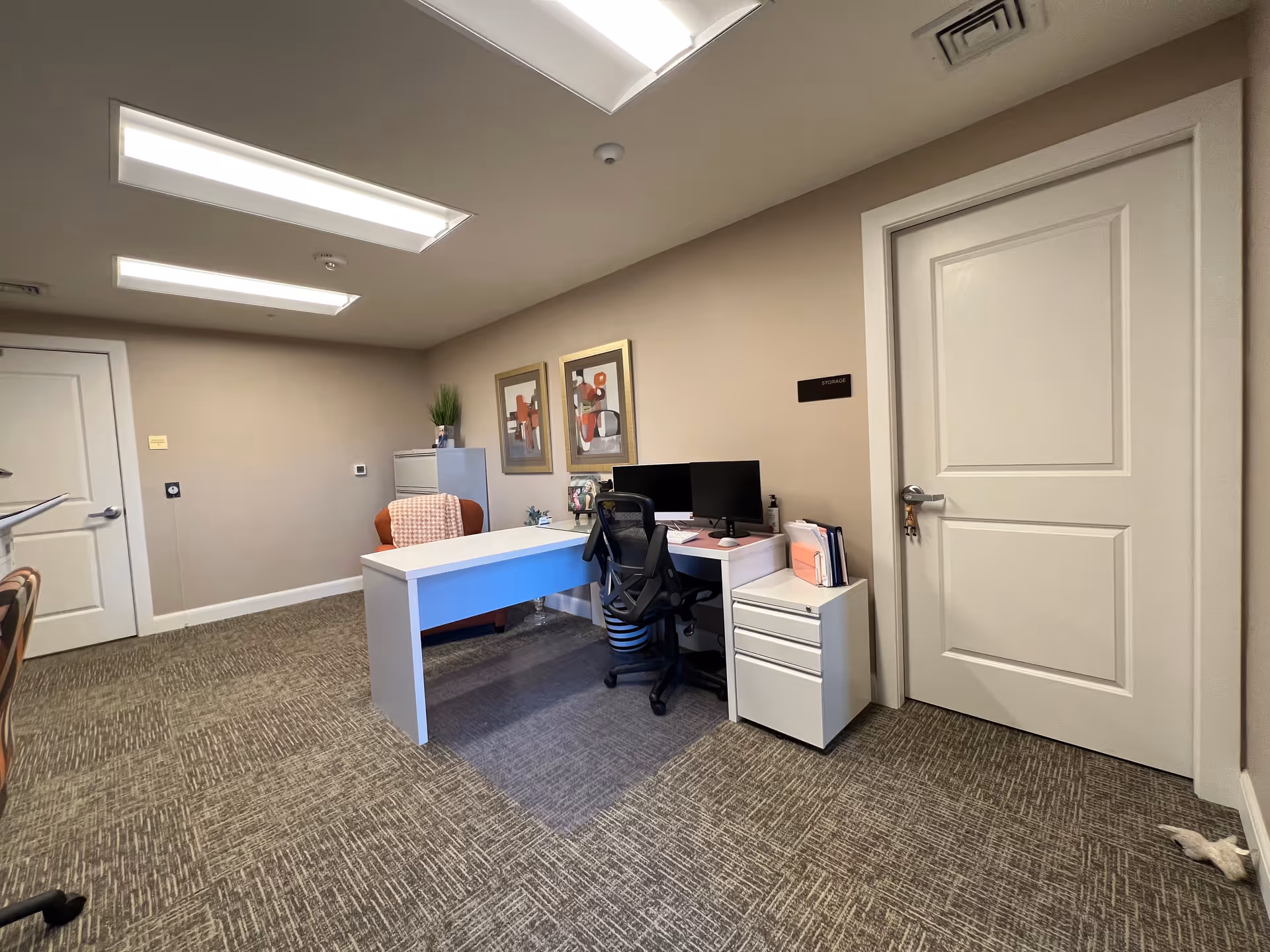 Office desk with computer, filing cabinet, and framed art in a neutral administrative room.