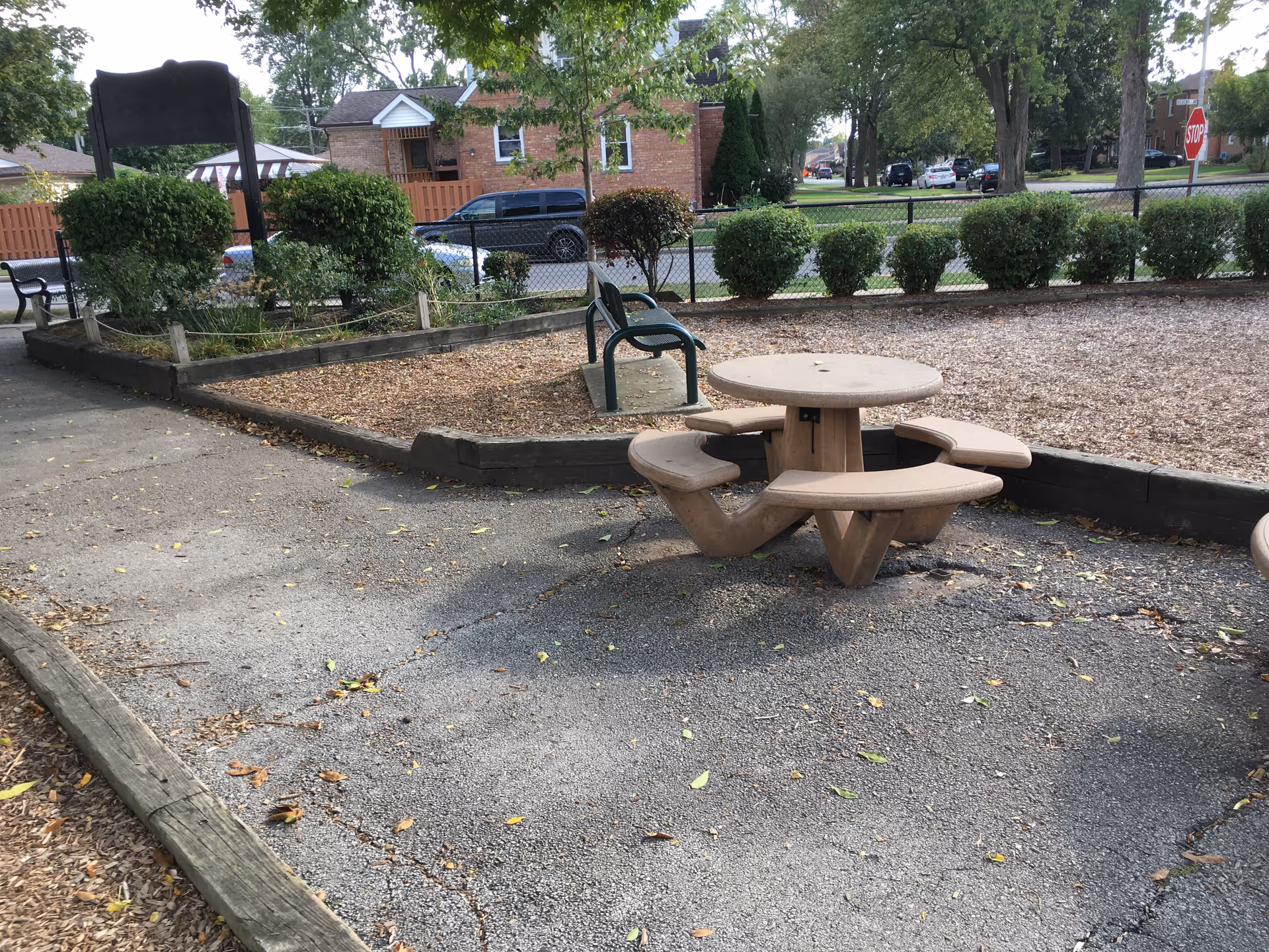 Outdoor courtyard with a round picnic table, mulch beds, shrubs, and a bench near a building.
