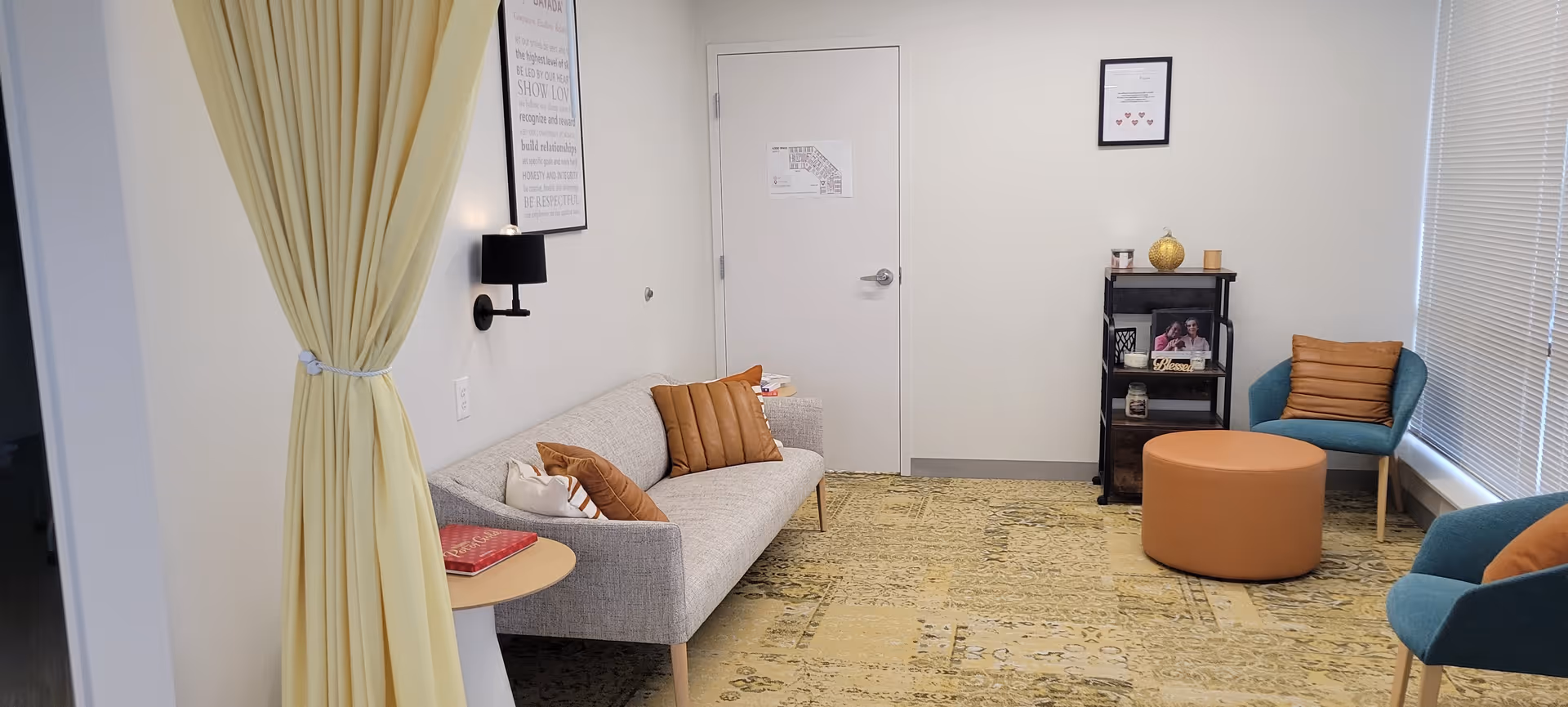 Bright senior living lounge with couch, chairs, ottoman, and bookshelf near a door.