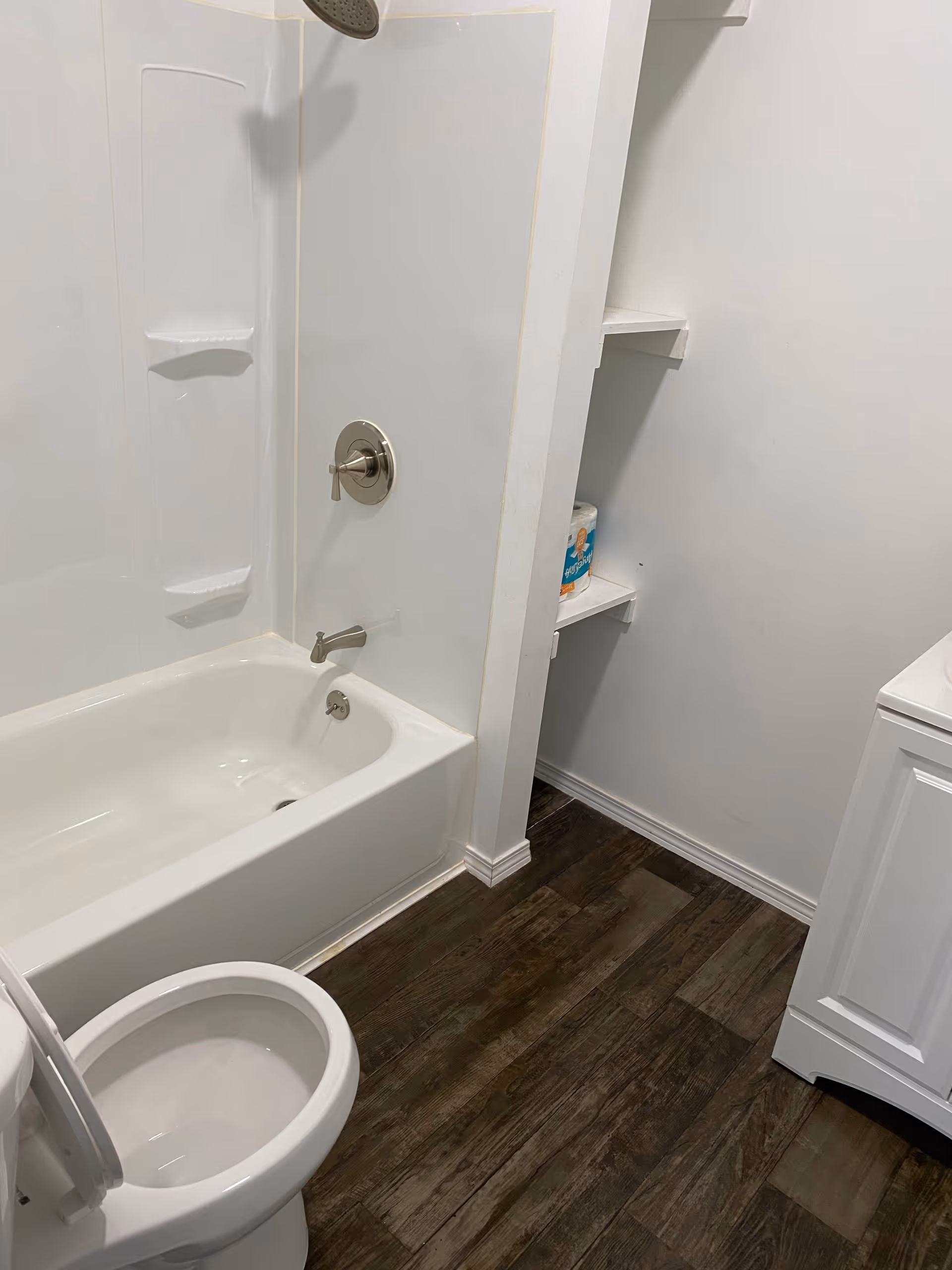 White bathroom with tub/shower, toilet, and shelving beside a vanity.