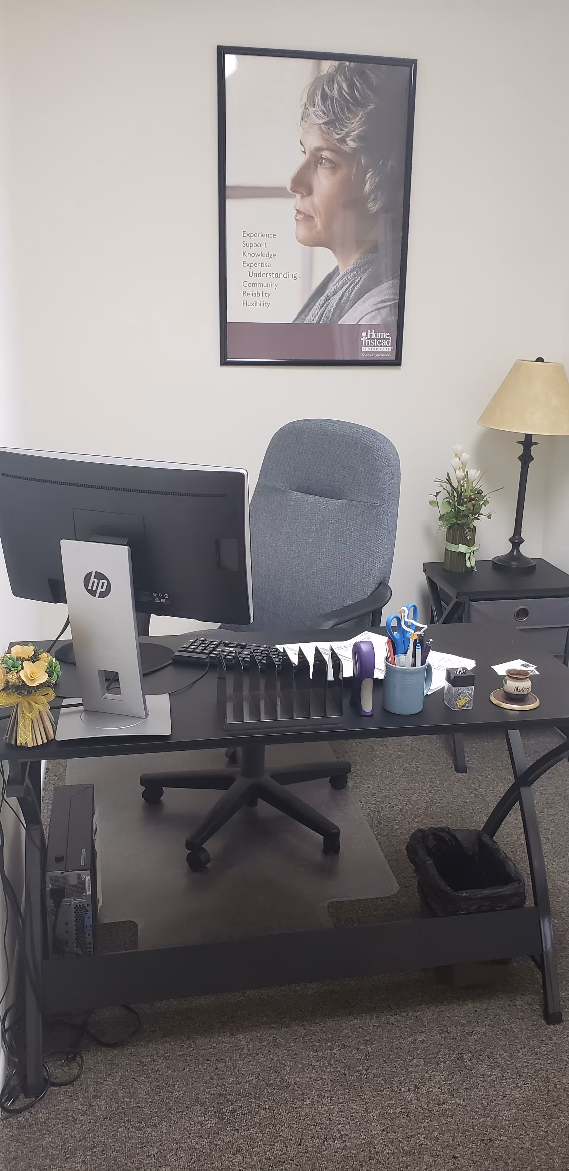 Office desk with computer, chair, supplies, and a framed poster on the wall at Home Instead.