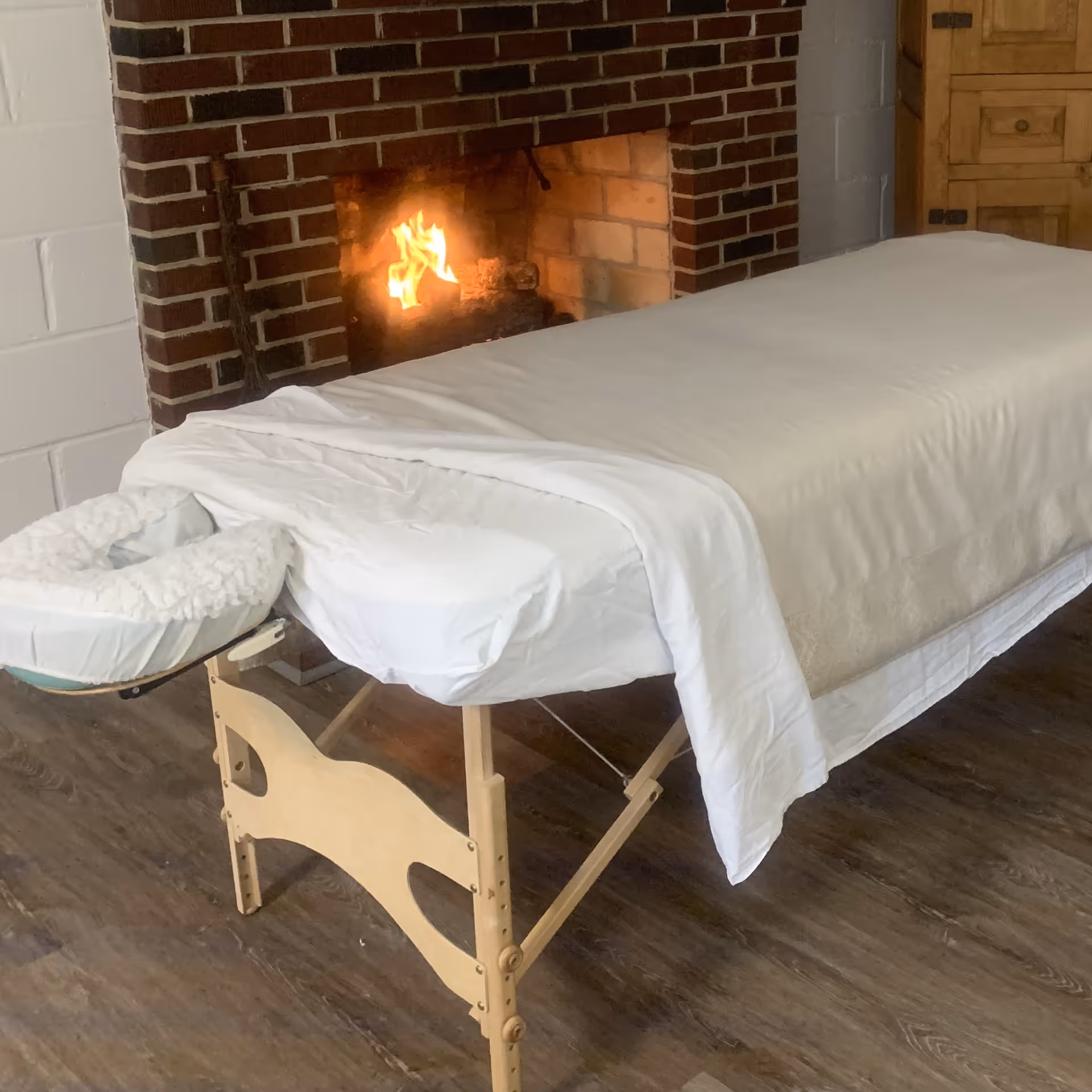 A massage table with white sheets beside a brick fireplace in a cozy room.