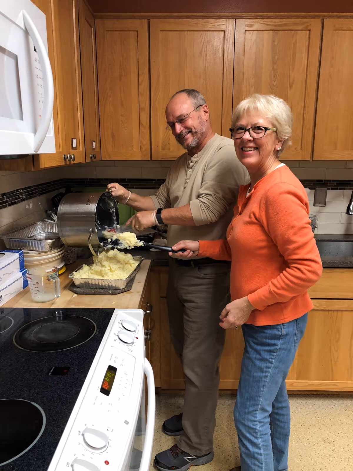 Two seniors cook mashed potatoes together in a cozy wooden kitchen, smiling at the camera.