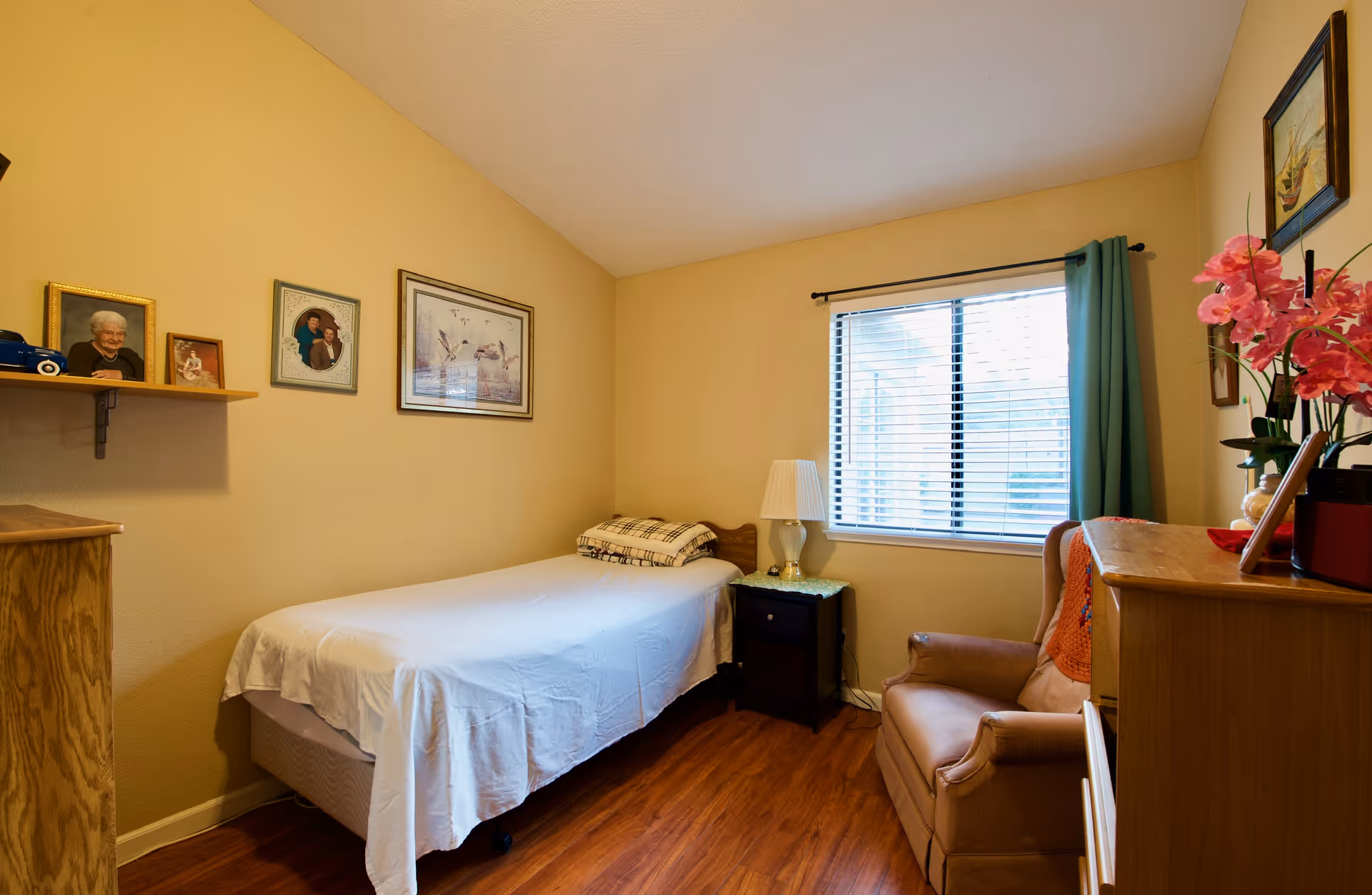 Small bedroom in a senior facility with bed, nightstand, chair, window, and photos on wall.