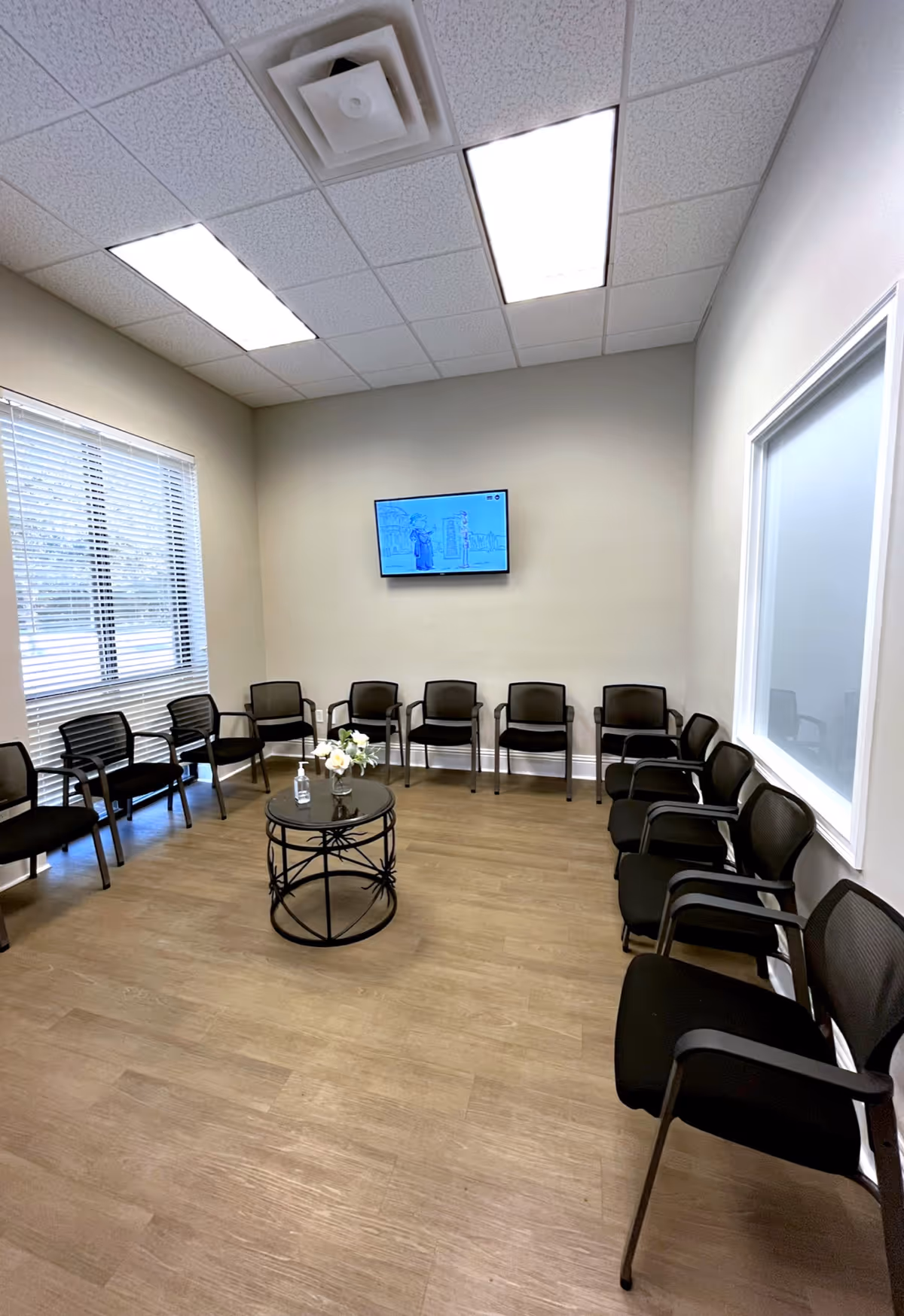 A waiting room with multiple black chairs around a round glass table with a flower vase.
