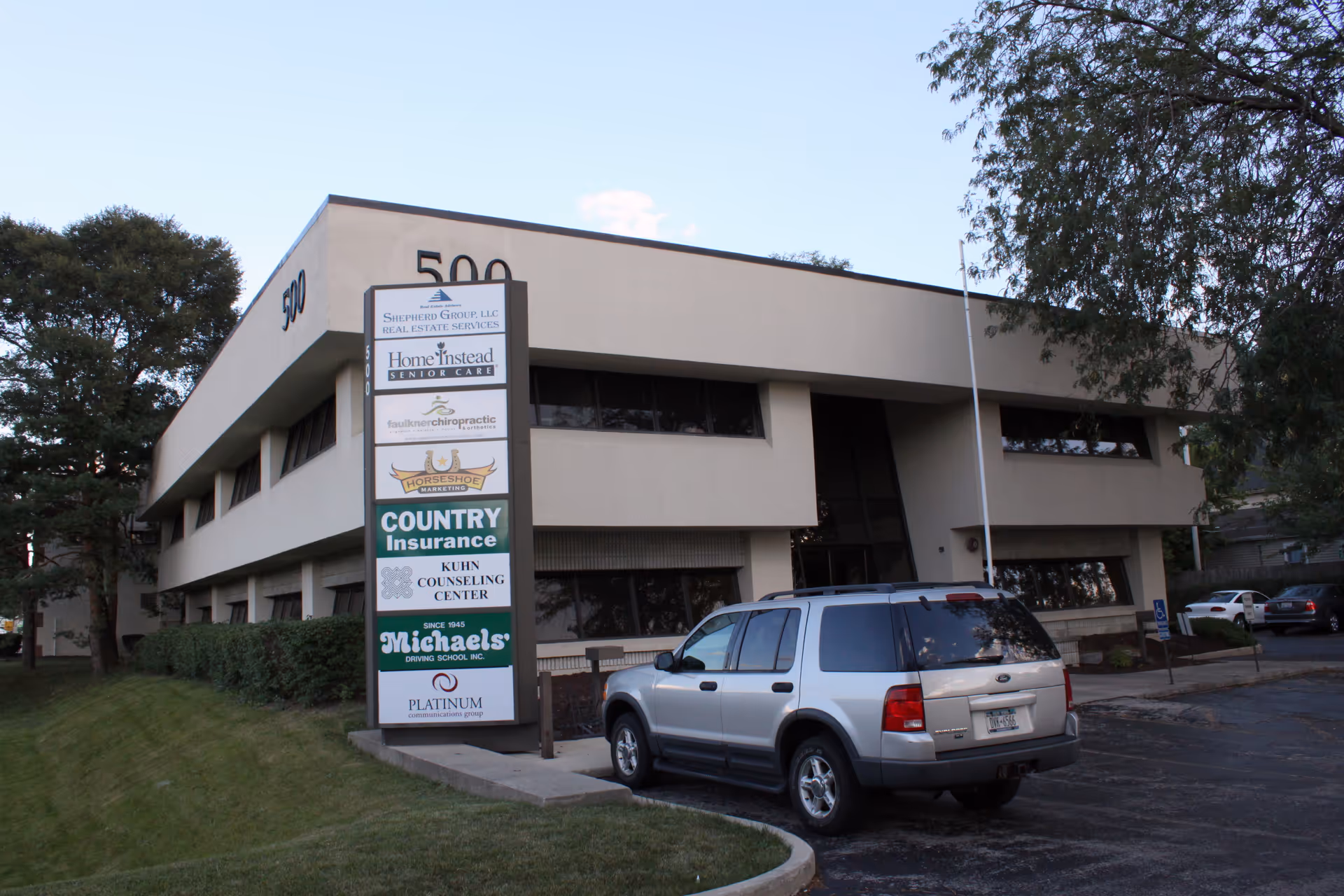Exterior view of a beige building with a tall sign and a parked SUV.