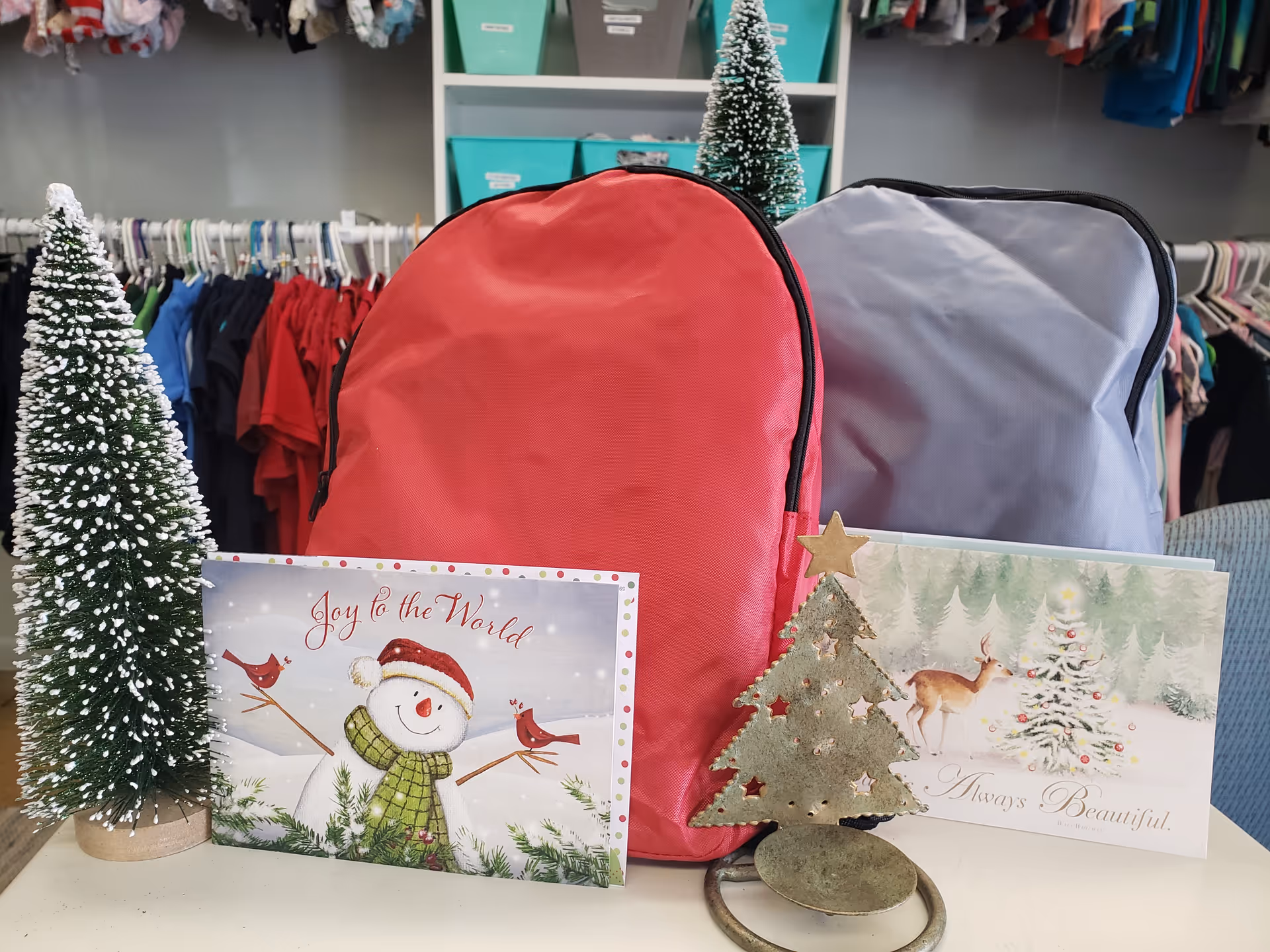 Red and gray backpacks on a table with Christmas cards and miniature trees.