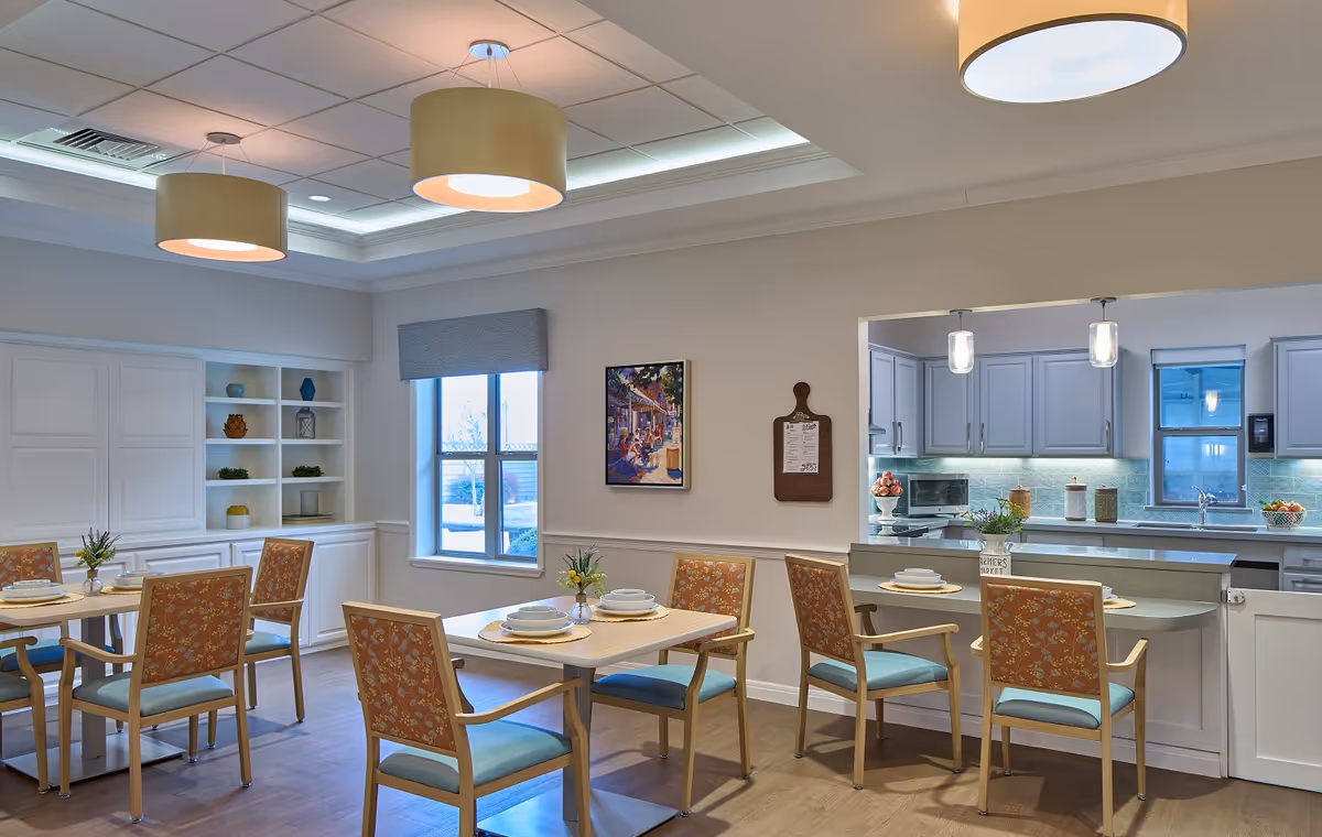 Bright dining room with multiple tables and chairs and a visible kitchen area in the background.