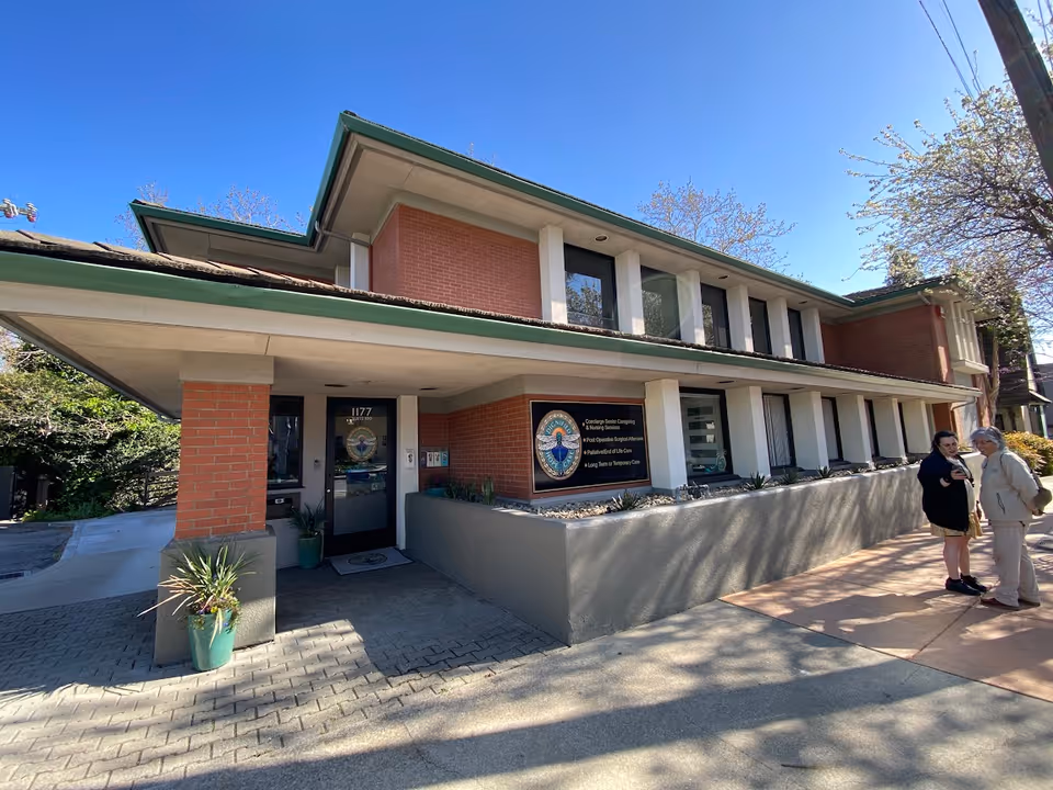 Two people stand outside a brick senior care facility entrance under a clear blue sky.