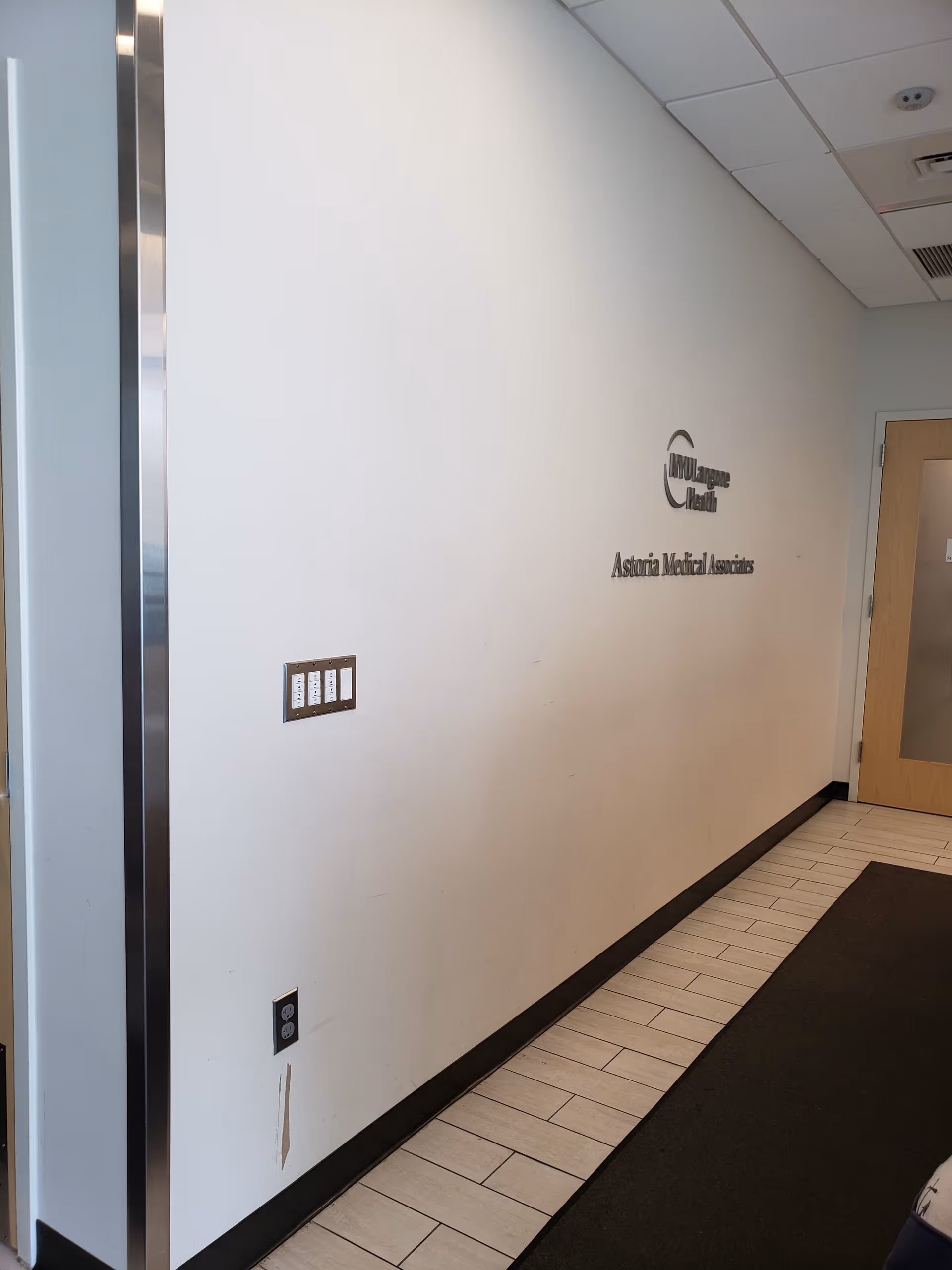 White interior hallway with wall-mounted NYU Langone Astoria Medical Associates sign and doorway.