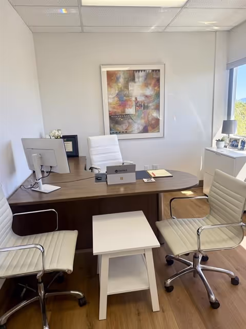 A modern office with white chairs, a wood desk, computer, and artwork.