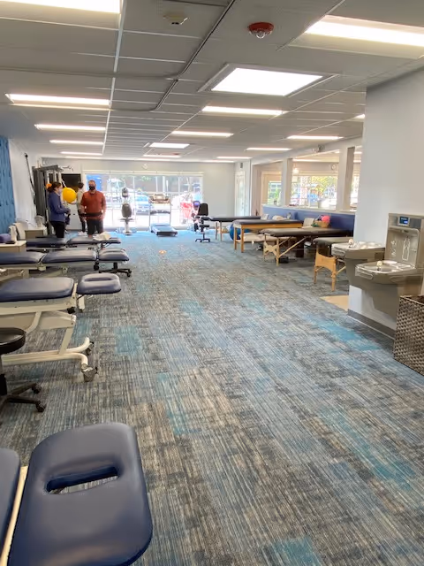 Open therapy clinic interior with treatment tables, chairs, and several people.