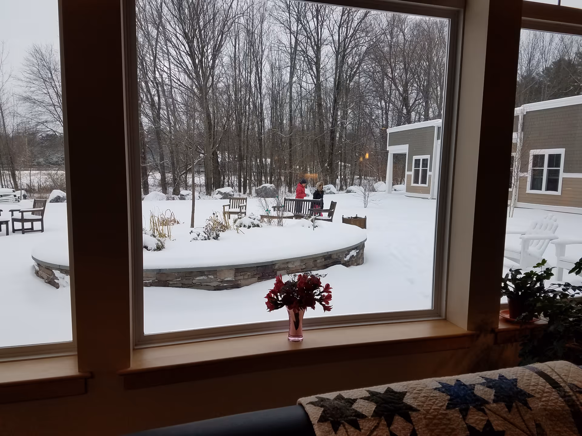 Snow-covered outdoor courtyard viewed through a window, with benches, planters, and distant figures.