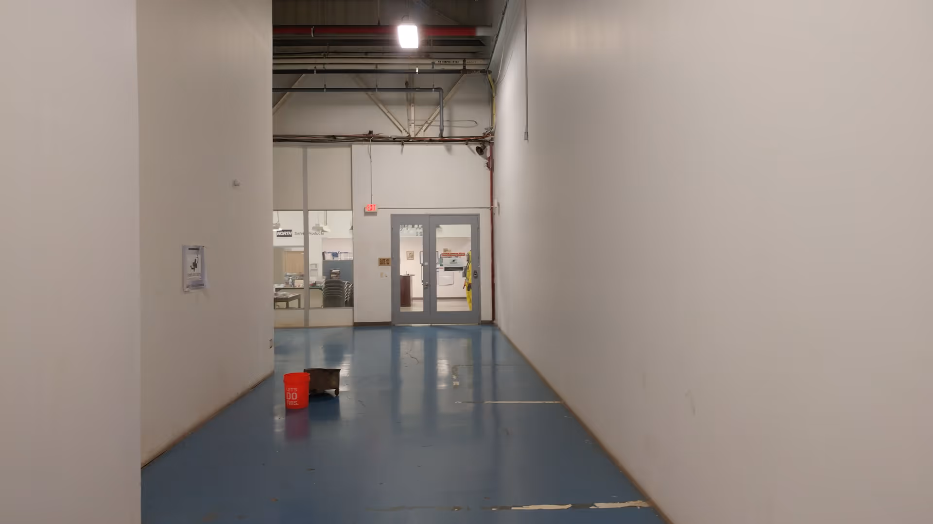 Long, empty hallway inside facility with blue floor, white walls, and a cleaning bucket.