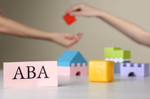 Two hands exchange a red block across a table with ABA therapy materials.