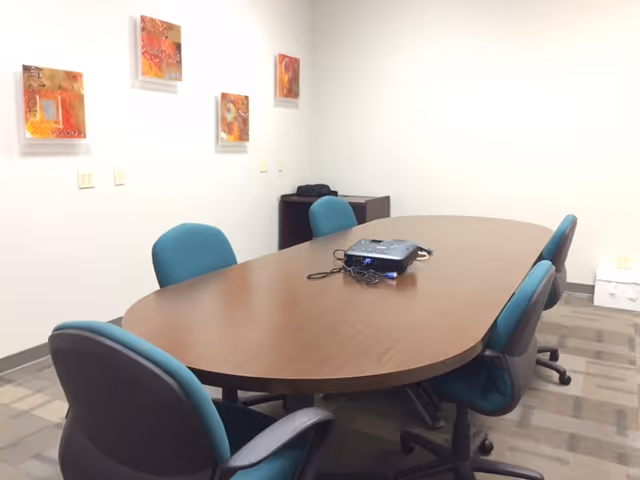 Oval wooden conference table with teal chairs in a bright meeting room; projector on the table.