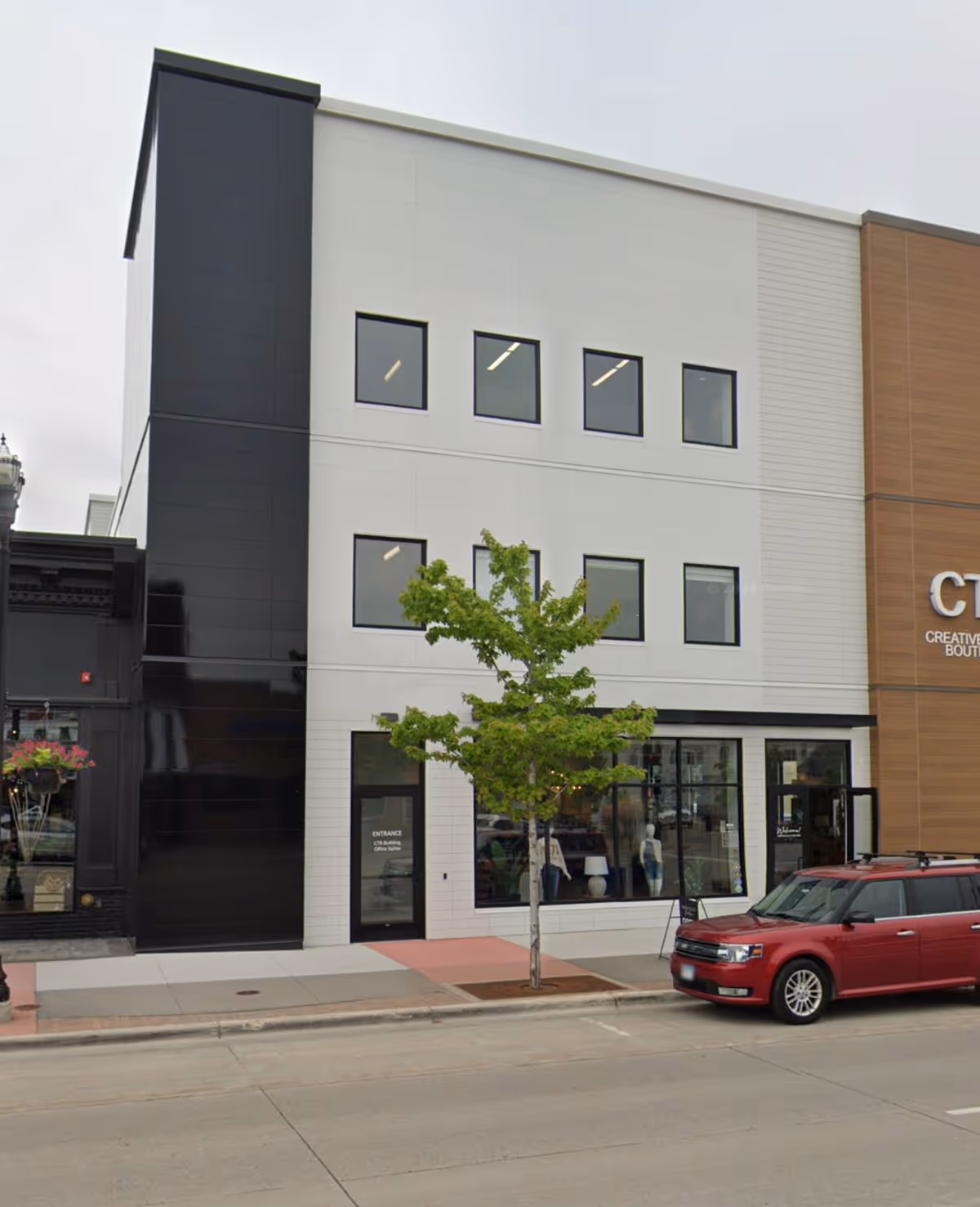 Modern three-story building front with large windows, a tree, and a parked red van.