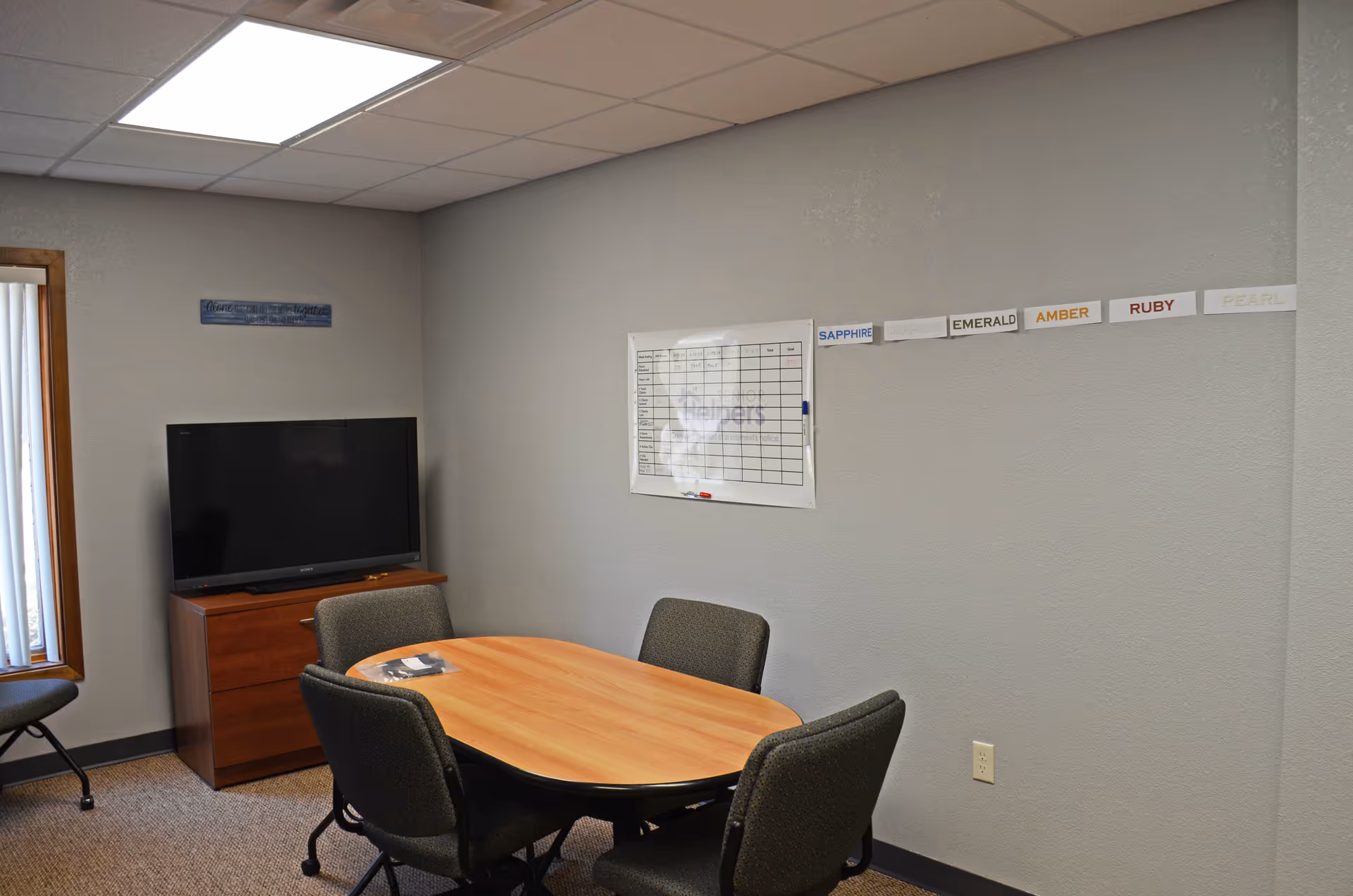 A small meeting room with a wooden table, chairs, a TV, and a wall chart.