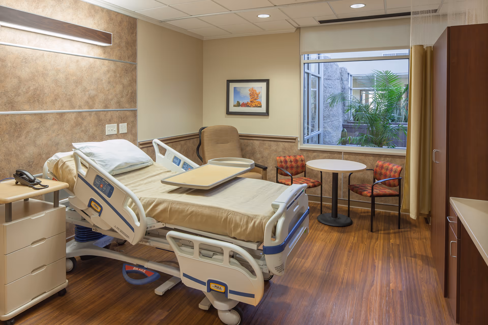Hospital-style patient bed in a clean room with table, chairs, and window.