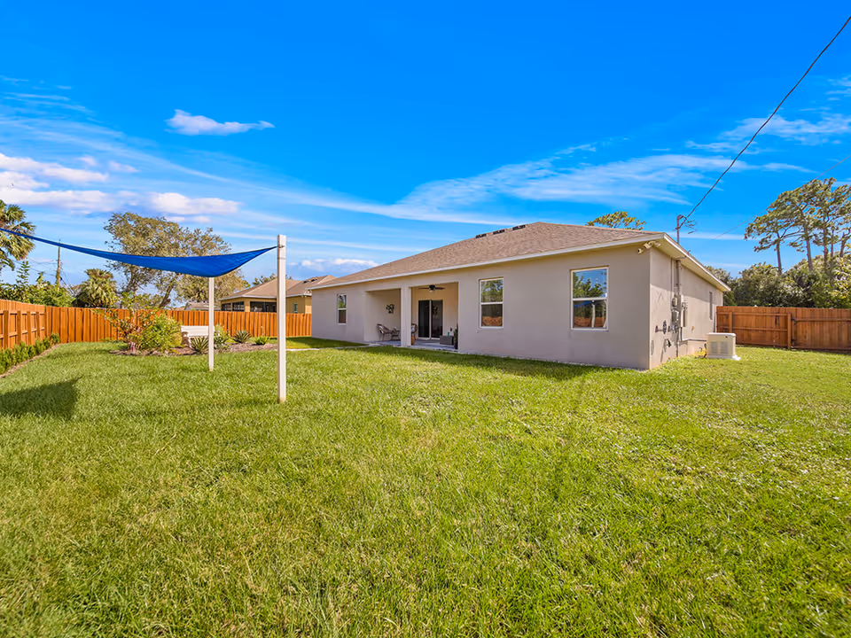 Backyard of a single-story home with a large grassy lawn, fence, and a blue shade sail.