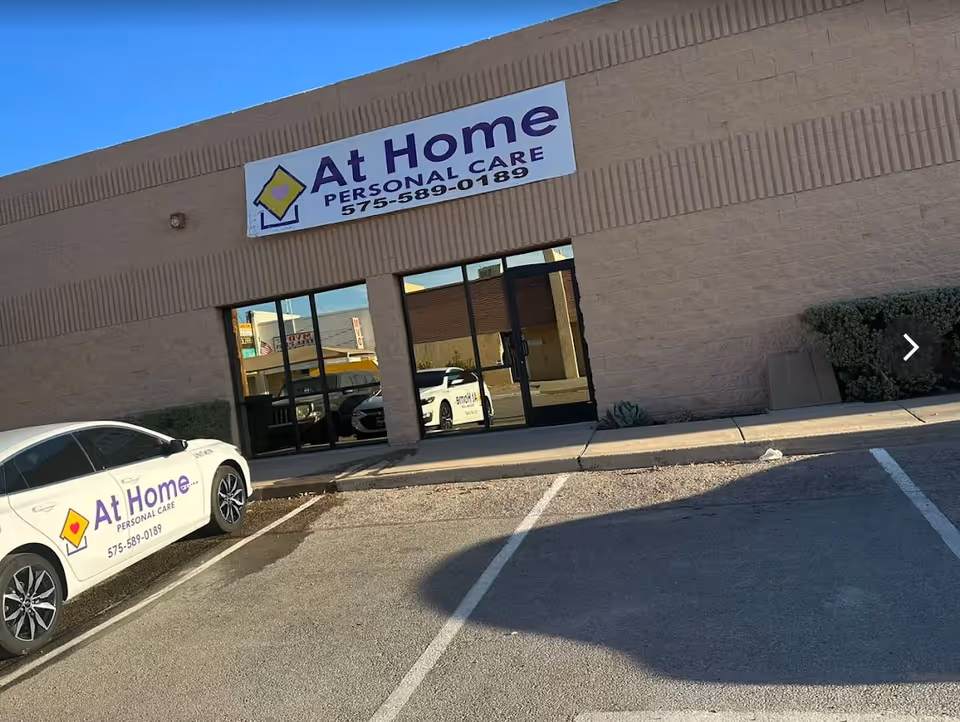 Exterior of At Home Personal Care building with sign, glass doors, and parked cars.
