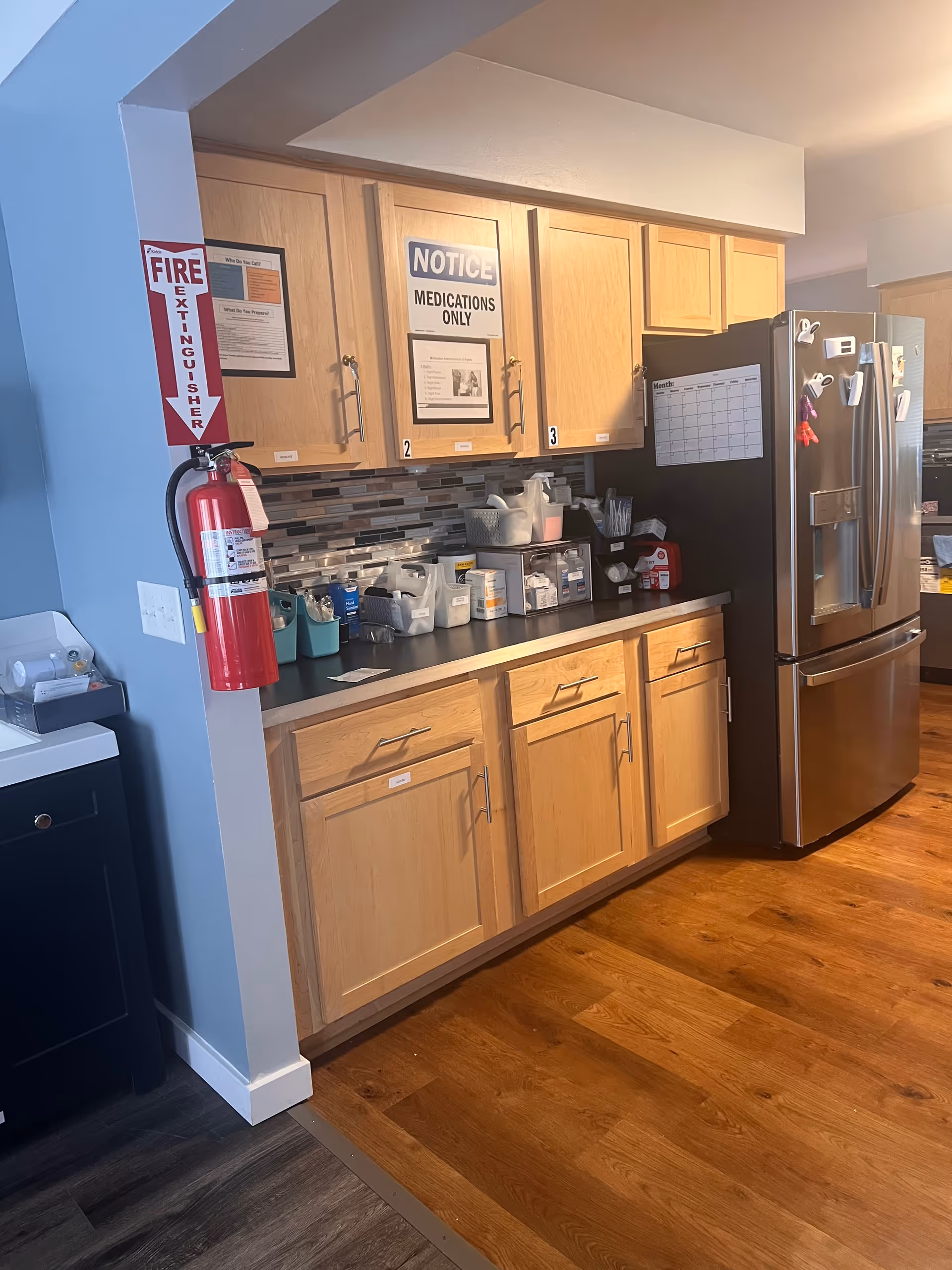 Wooden-cabinet kitchen area with counter, supplies, and stainless refrigerator in a senior facility.