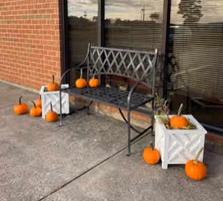 Outdoor brick wall scene with a bench, pumpkins, and white planter crates on the sidewalk.