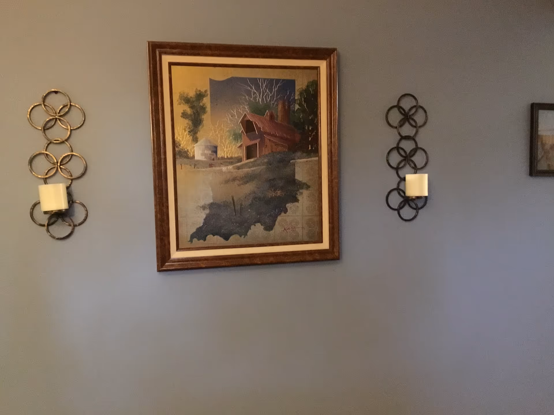 Gray interior wall with a framed rural landscape painting and decorative circular wall sconces.