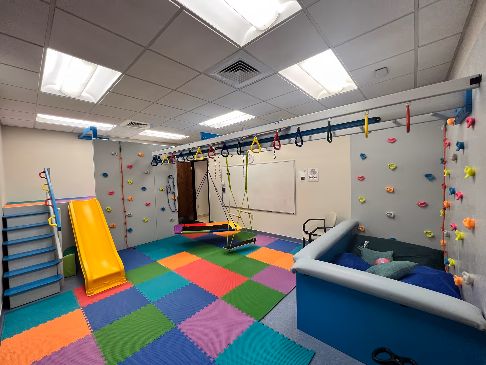 Colorful indoor therapy playroom with climbing walls, soft mats, a slide, and a cushioned seating area.