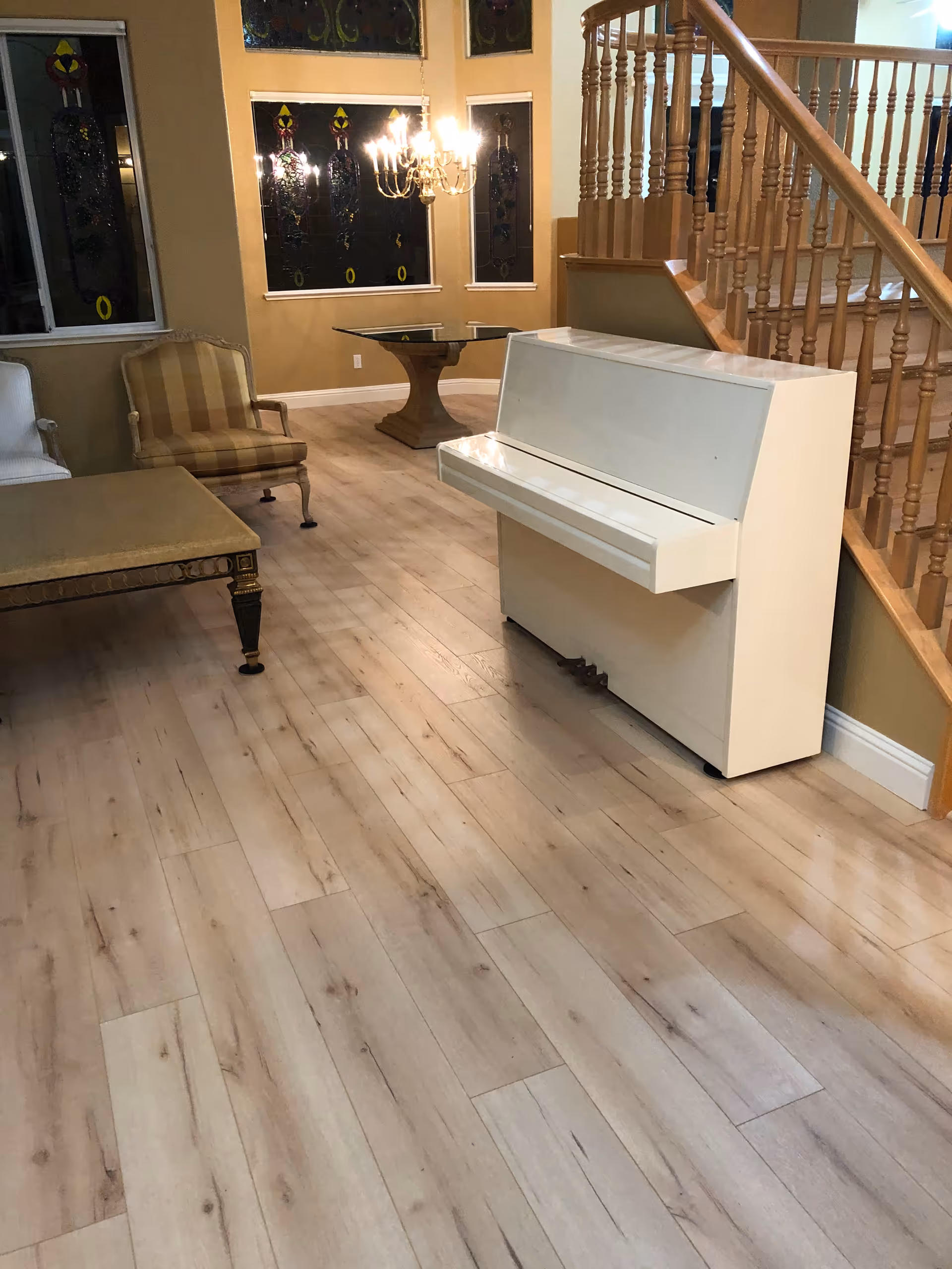 Interior living room with a white upright piano, chandelier, seating, and a wooden staircase.