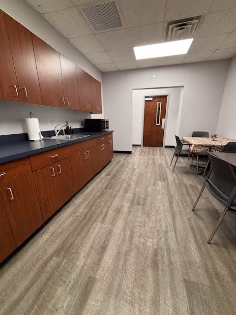 Long senior living kitchenette with wooden cabinets, sink, microwave, and a dining table with chairs.