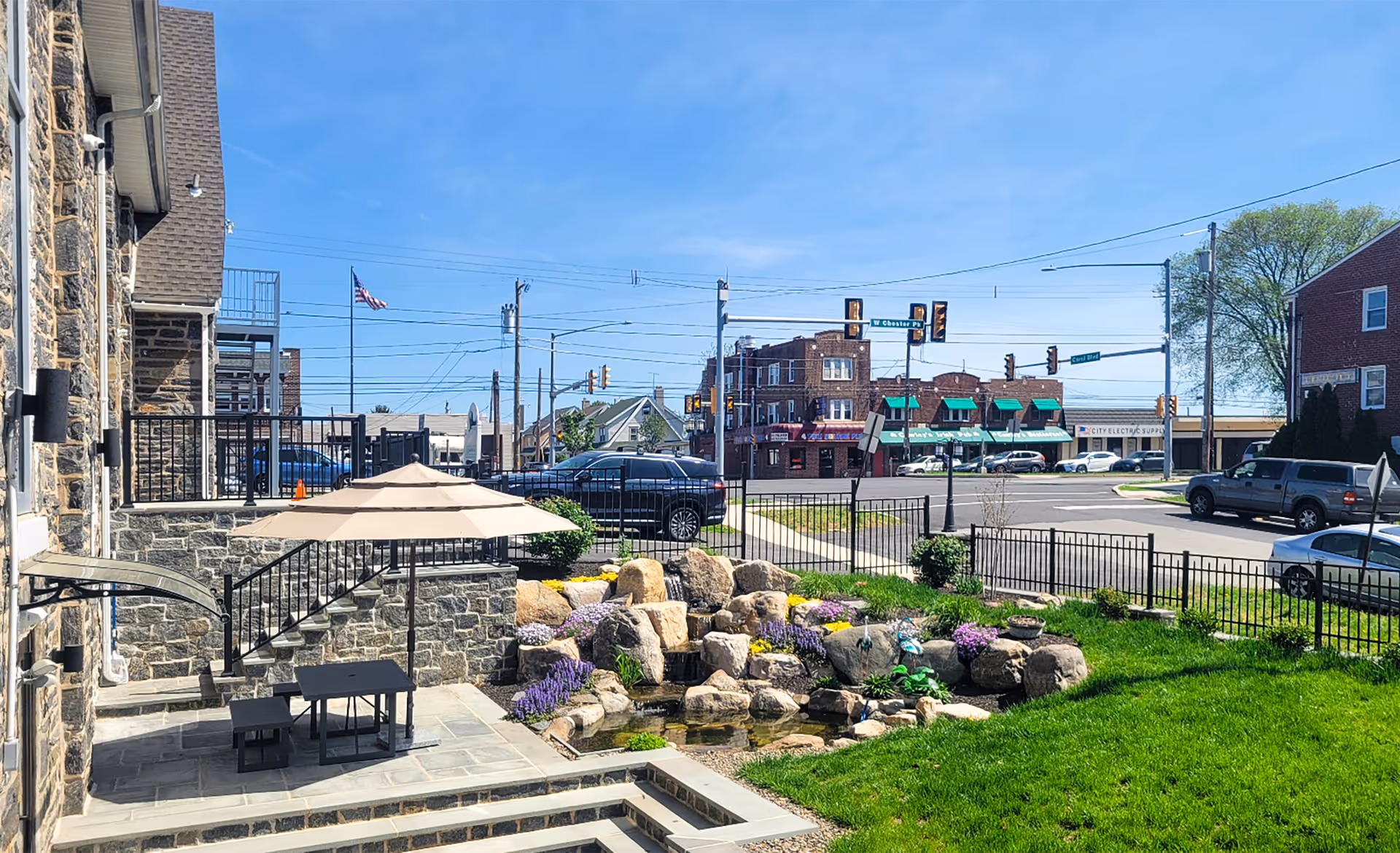 Outdoor patio with table, umbrella, rock garden, and stone building overlook a street.