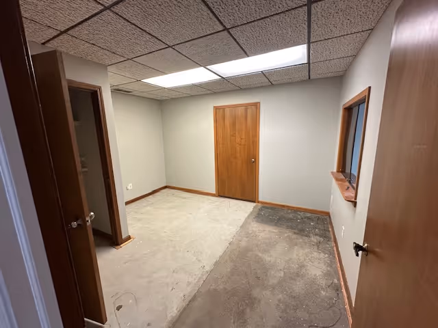 A small empty interior room with a drop ceiling, doors, window, and uneven flooring.