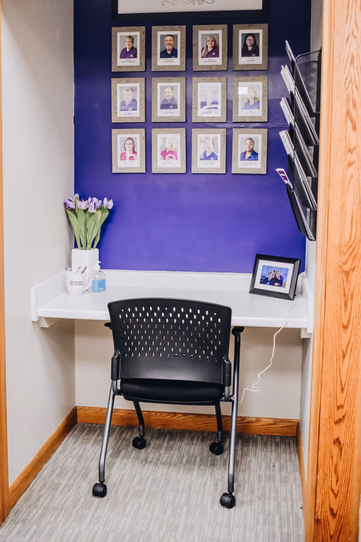 A small desk area with a purple wall, framed photos, and a black chair.