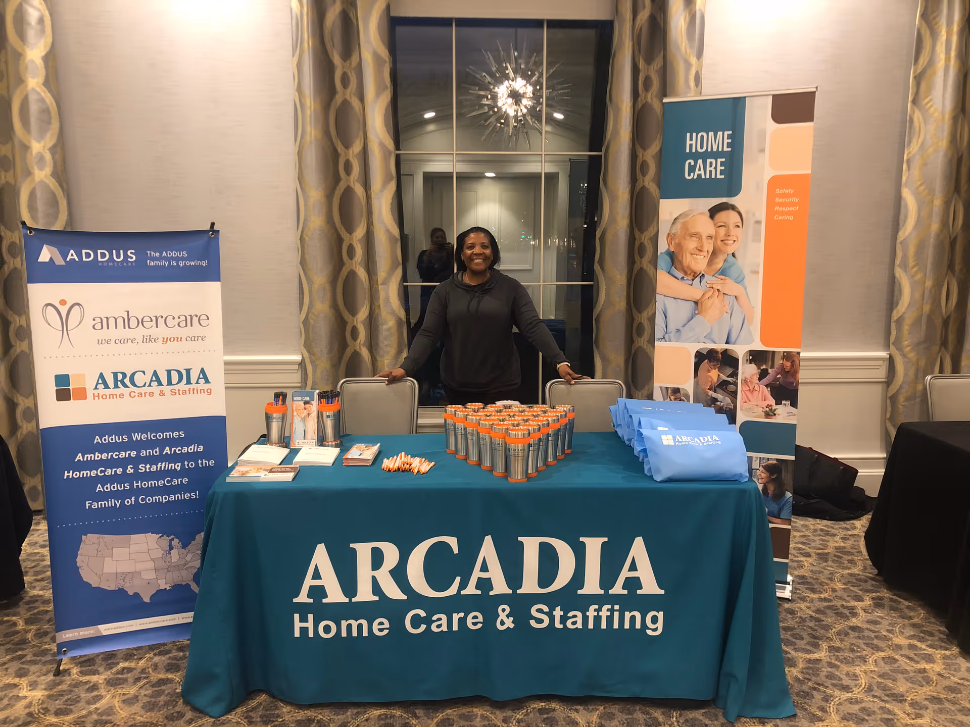 A person stands behind a teal Arcadia Home Care booth at an indoor event.