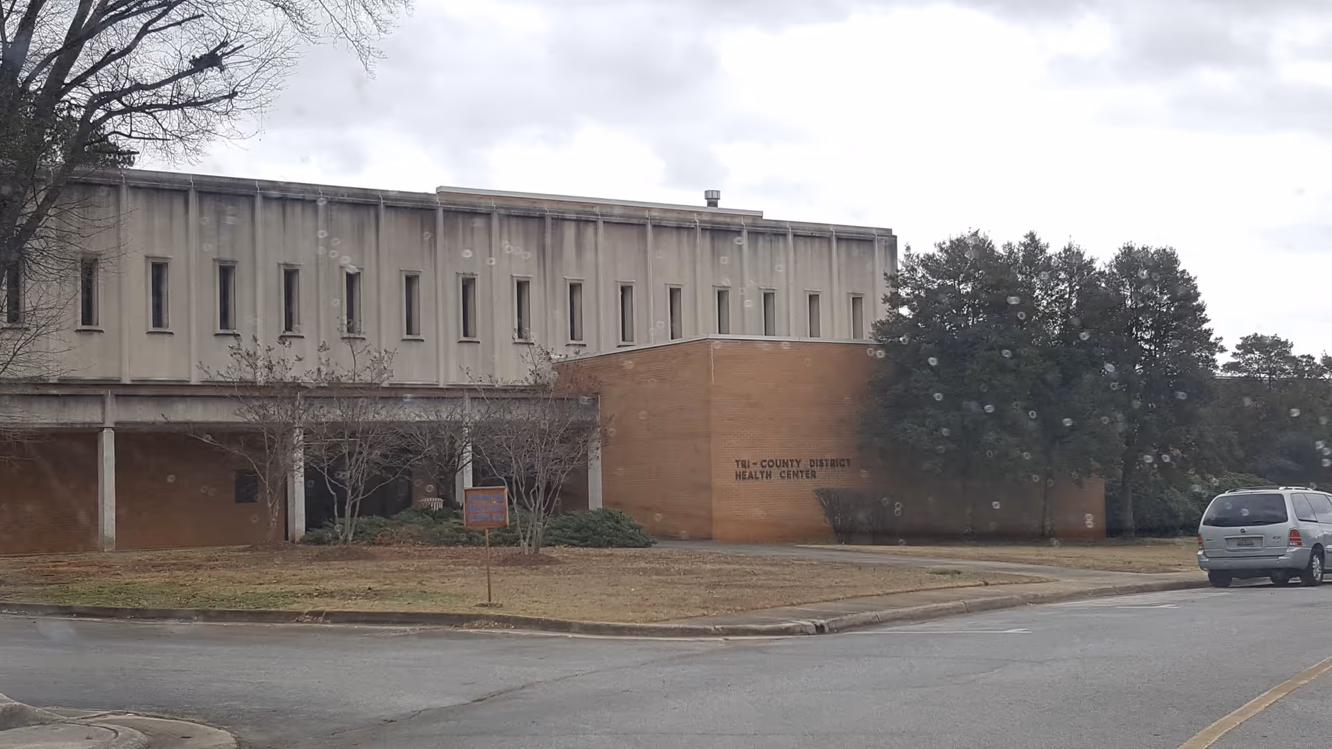 Exterior view of a large beige health department building with a parked minivan nearby.