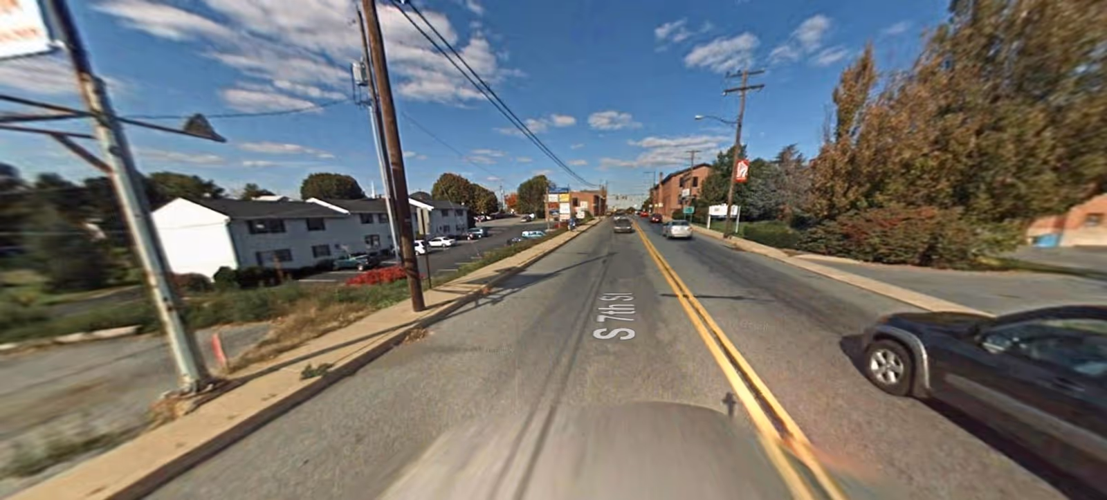 A street view capturing a sunny avenue with sidewalks, utility poles, trees, and parked cars.