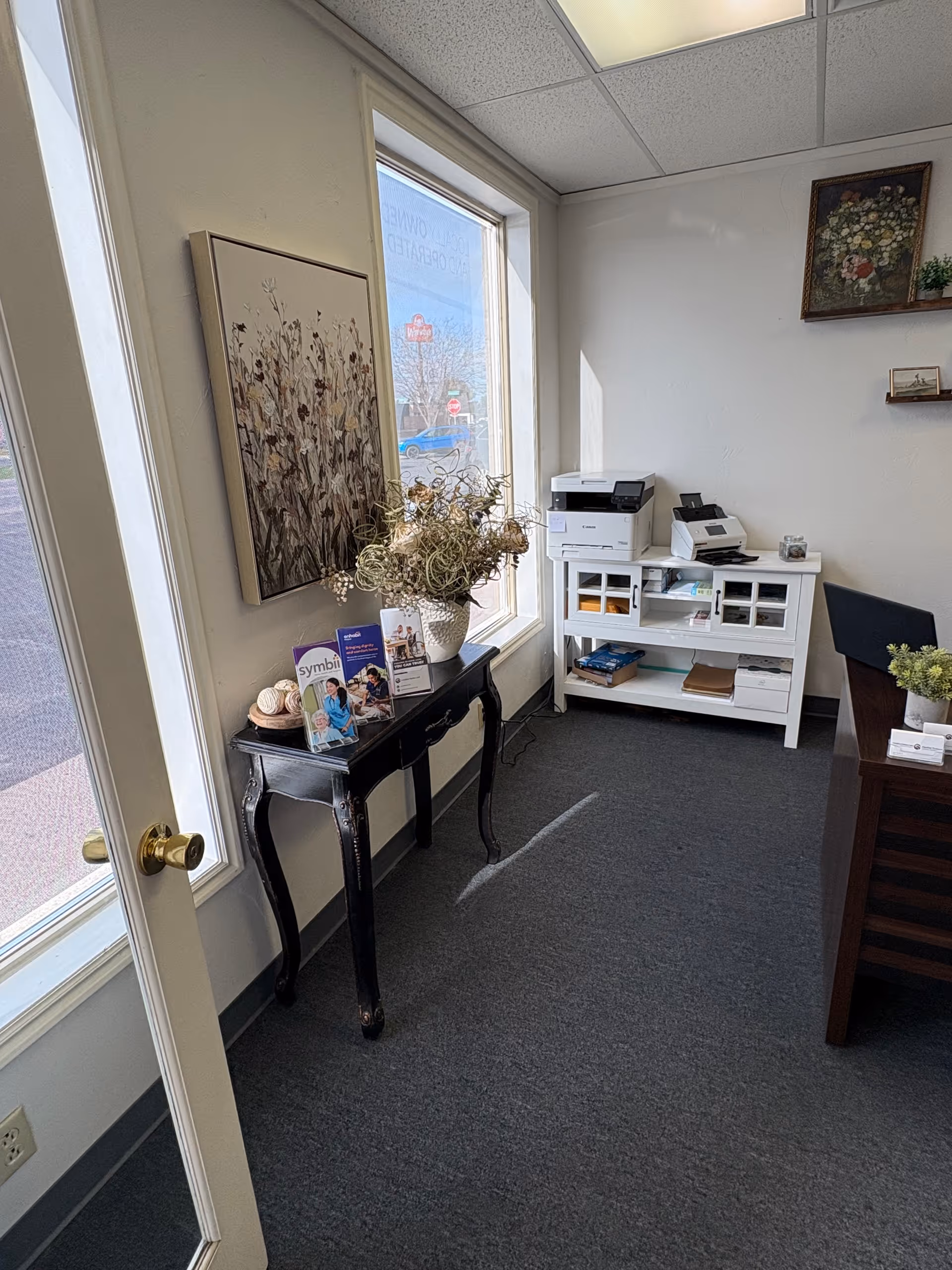 Interior lobby with window, black console table, brochures, dried flowers, and a white printer cabinet.