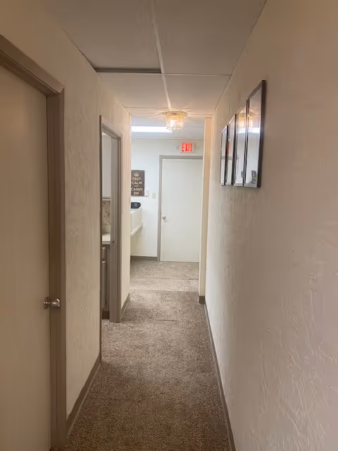 Long carpeted hallway in a senior living facility with doors, framed pictures, and an exit sign.