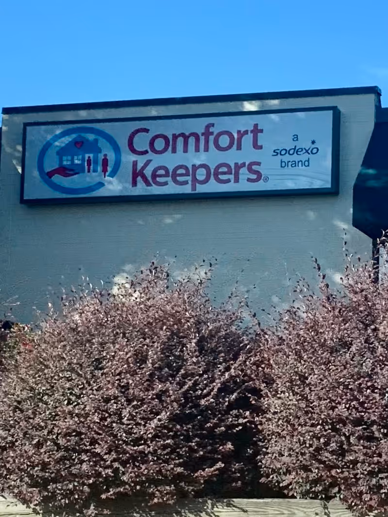Exterior view of a Comfort Keepers sign on a building, with bushes in foreground