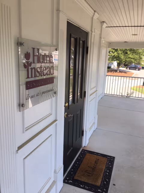 Exterior view of Home Instead entrance with sign, black door, and covered porch.