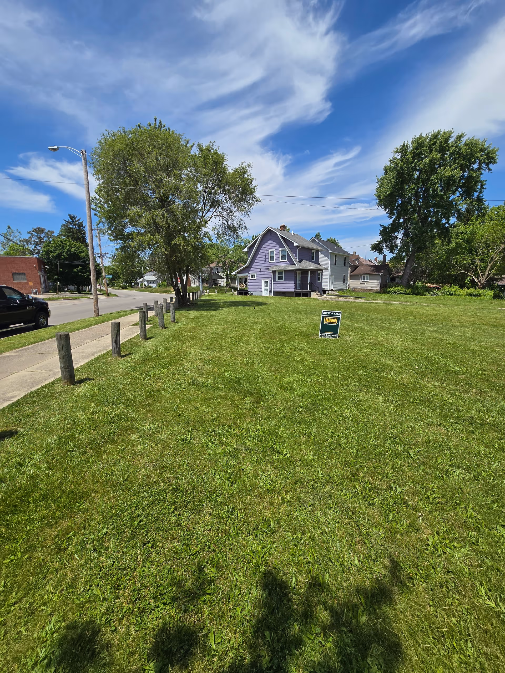 Green lawn in front of a purple house with a For Sale sign and trees.